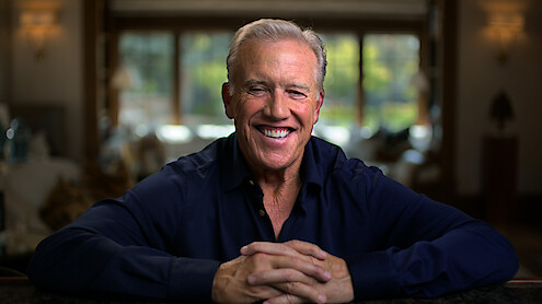 Smiling man with gray hair wearing a dark shirt sits indoors with hands folded, relaxed, in a warmly lit, blurred living room with large windows and natural light in the background.