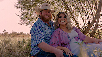 Smiling couple sitting together outdoors in a grassy field, with the man wearing a cowboy hat and patterned shirt, and the woman in a light dress and floral shawl. Sunlit trees and warm, natural light in the background.