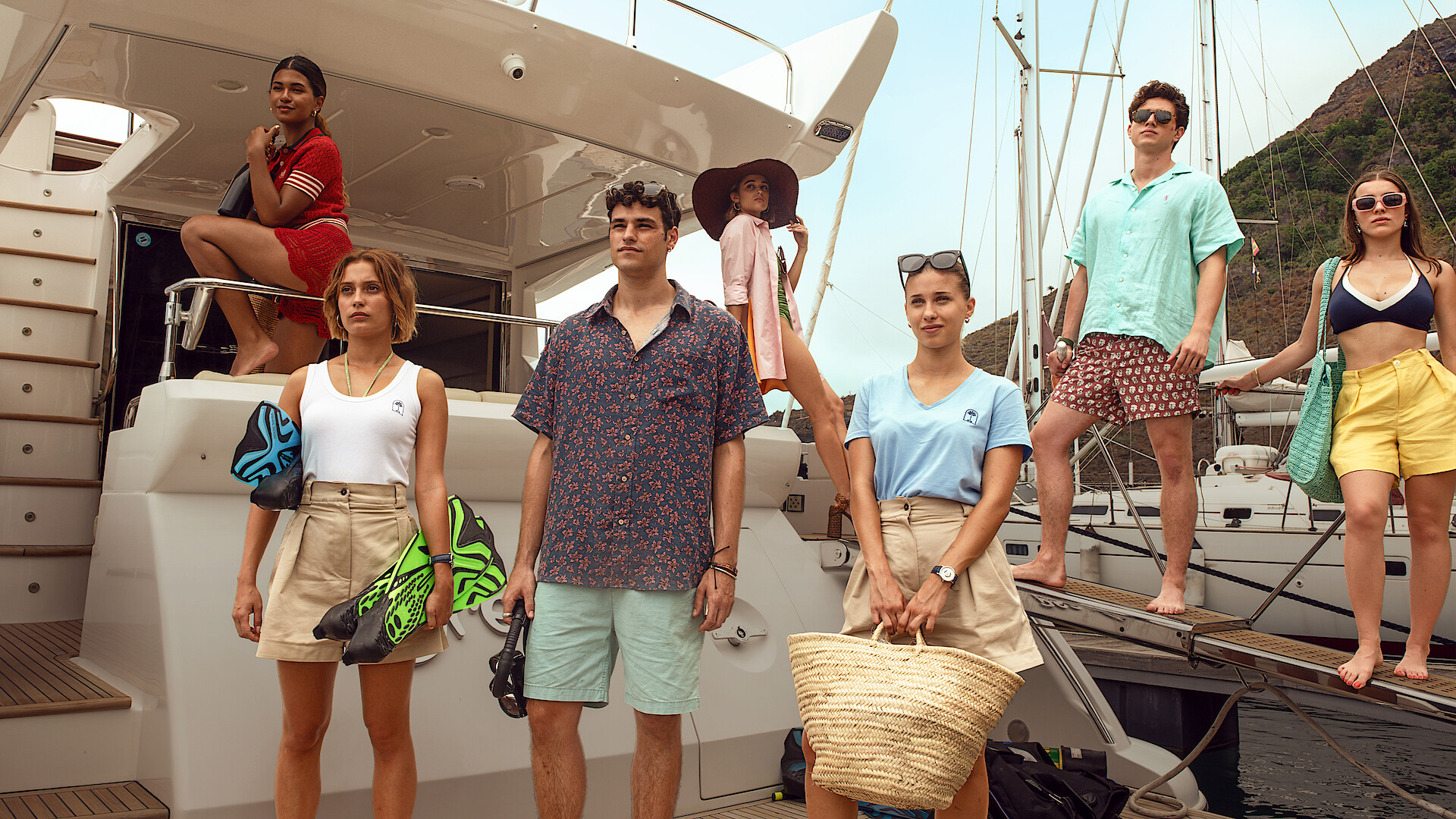 Group of young adults in summer outfits on a docked yacht, posing confidently. The marina background shows sailboats and a hillside, suggesting a relaxed, upscale vacation setting with sunny weather.