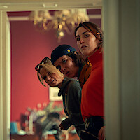 Four silhouetted figures stand in front of a burning wooden house at night, surrounded by trees, as flames engulf the structure, creating a tense and dramatic atmosphere.