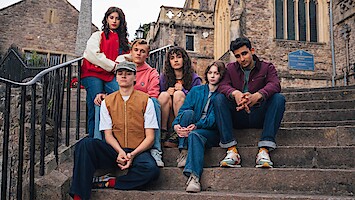 Six young adults sit on stone steps outside an old stone building with arched windows, likely a church or historic structure, dressed in casual, colorful clothing and posing as a group.