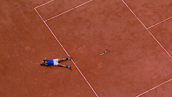 A tennis player lies on a clay court next to a tennis racquet, possibly celebrating or exhausted, viewed from above. The scene captures the lines and earthy texture of the tennis court.