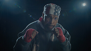 A male boxer wearing red hand wraps and a USA flag bandana poses in a fighting stance under dramatic lighting in a dark gym or training environment.
