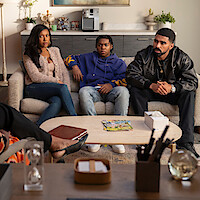 Three people sit on a sofa in a modern living room, appearing serious or thoughtful, while another person’s legs are visible in the foreground, suggesting a discussion or therapy session. The room is decorated with plants and books.