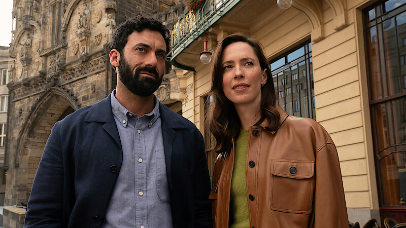 A man and woman stand outdoors in a city setting, with historic stone building details and modern architecture visible behind them. Both appear thoughtful, dressed in casual jackets, under daylight.