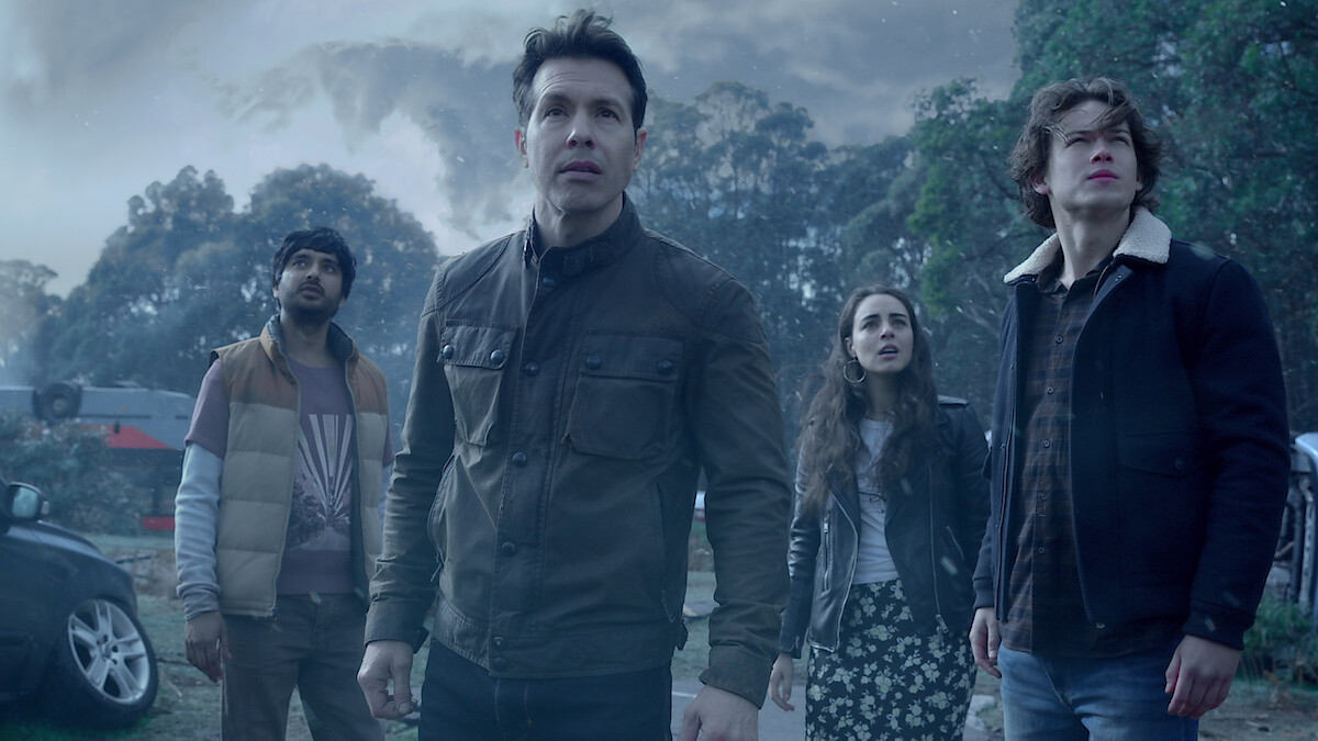 Four people standing outdoors in a tense atmosphere, surrounded by cars and trees under a stormy, cloudy sky, all looking up with concerned expressions.