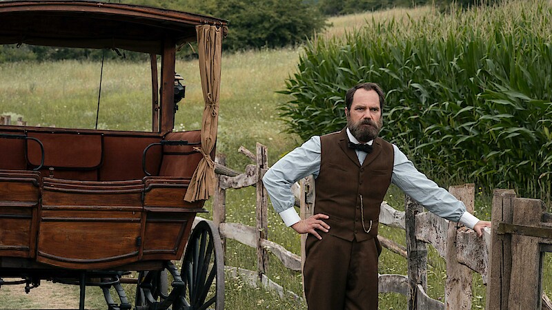 A man in period clothing stands by a wooden fence next to an old carriage in a rural setting with green cornfields and meadows in the background.