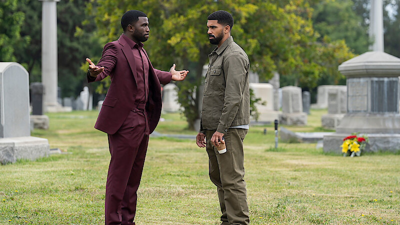 Two men stand in a cemetery, surrounded by gravestones and greenery; one man in a burgundy suit gestures with open arms while the other, in casual clothing, holds a coffee cup and listens.