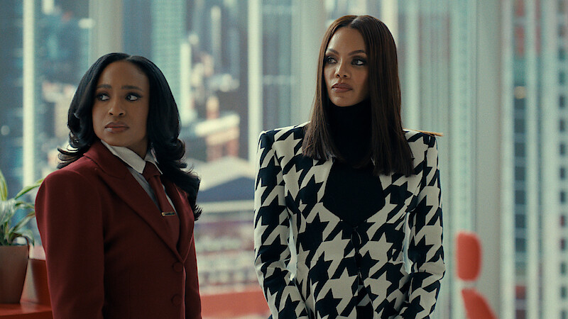 Two women stand in a modern high-rise office with large windows showing a cityscape; one wears a red suit, the other a bold black and white patterned jacket, both looking serious and professional.