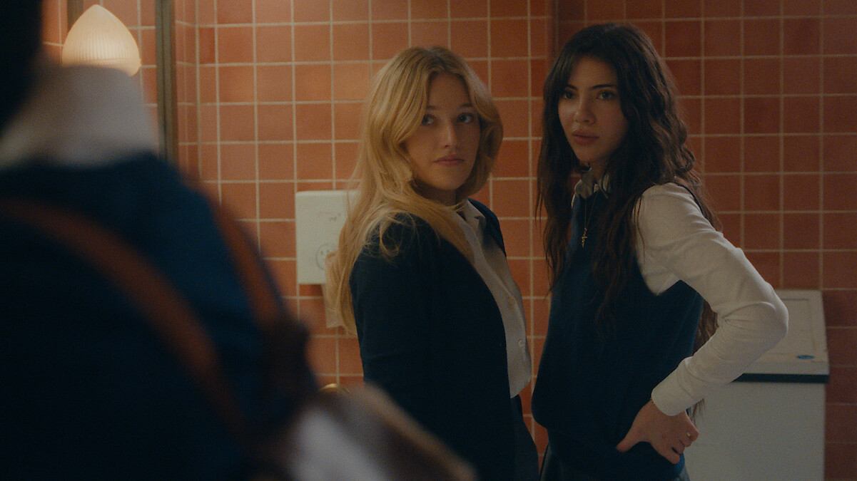 Two young women in school uniforms stand in a bathroom with pink tiled walls, looking toward someone entering. The setting looks like a school. One has blonde hair, the other has long dark hair, and both appear surprised or curious.