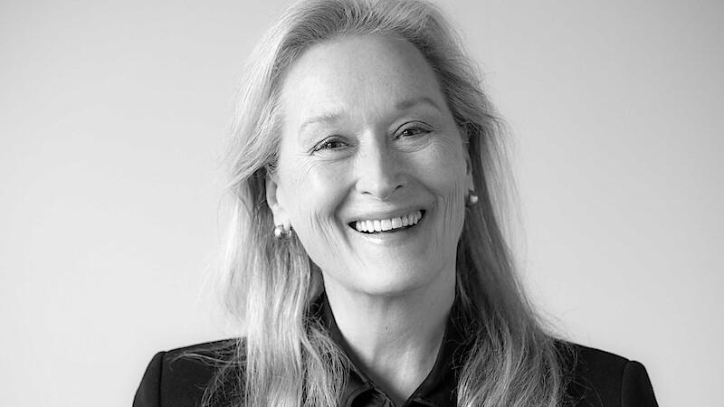 Smiling woman with long, light hair wearing a dark top, facing camera in a well-lit, plain indoor setting with a neutral background.
