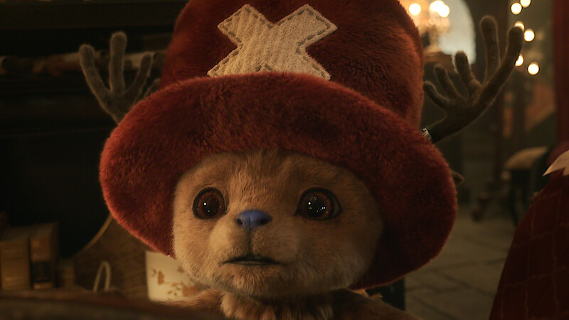 Tony Tony Chopper, a cute, furry character with antlers and a large red hat marked with a white X sits indoors in a dimly lit, cozy room with books and warm lighting in the background, looking surprised or curious.