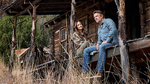 Man and woman sitting on porch of rustic wooden cabin in nature, surrounded by tall dry grass and trees, sharing a quiet moment outdoors.