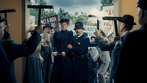 A woman in historical clothing is escorted by a uniformed officer through a crowd of protesters holding signs, squeegees, and newspapers in an outdoor setting during daylight.