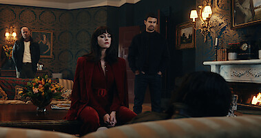 Kaya Scodelario as Susie Glass and Theo James as Eddie Horniman question someone seated on a couch in an ornate room in season 1 of 'The Gentlemen'