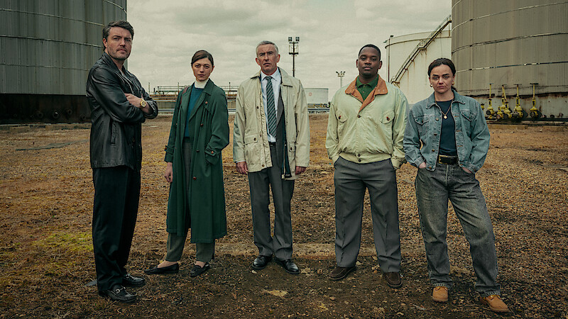 Five people stand in a gritty industrial outdoor setting between large storage tanks, dressed in various jackets and coats, facing the camera on a cloudy day, suggesting a serious or dramatic atmosphere.