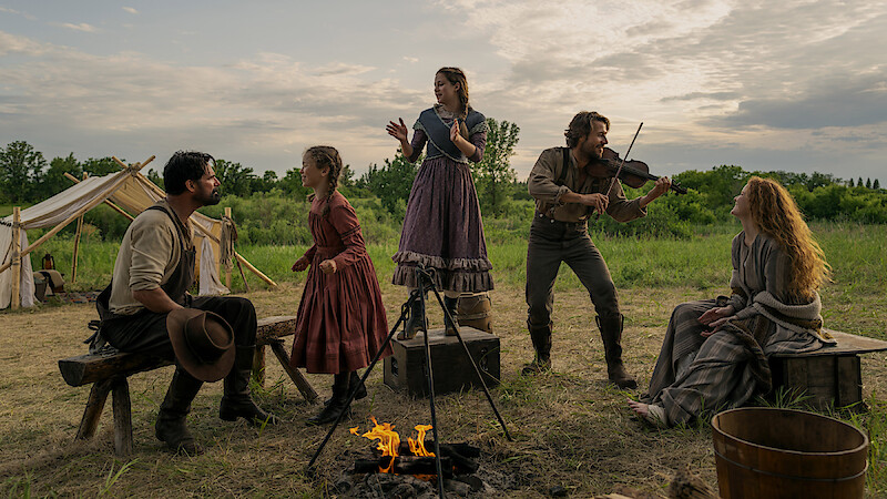 Five people in pioneer-era clothing gather around a campfire in an open field with tents. One plays a violin, others sing or listen, creating a lively, outdoor, historical family campsite scene at sunset.