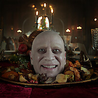 A smiling bald man’s head appears on a platter surrounded by roasted vegetables, set on an elegant dining table with ornate glasses and a dimly lit, candlelit gothic banquet hall in the background.