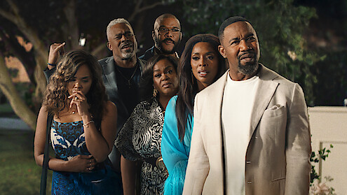Six adults stand outdoors at night, dressed in formal and semi-formal attire, looking intently in the same direction with serious and curious expressions; trees and garden setting in the background.
