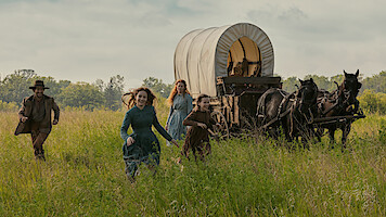 Four people in pioneer-era clothing run through a grassy field beside a covered wagon pulled by two horses, with trees and sky in the background, evoking a historical or frontier setting.
