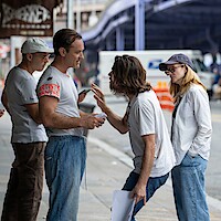 Four adults in casual clothing talking on a city street; one man gestures while speaking to another, with two others listening attentively. Urban setting with sidewalk, construction barriers, and vehicles in the background.