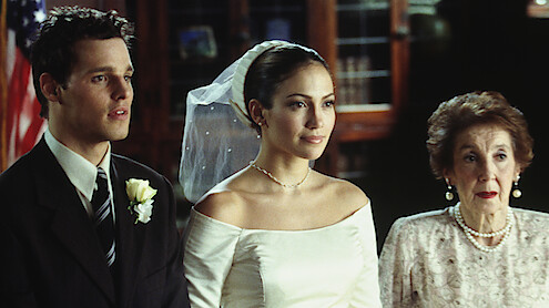 A bride in a white dress and veil stands between a man in a suit with a boutonniere and an elderly woman in pearls, indoors with bookshelves and an American flag in the background.
