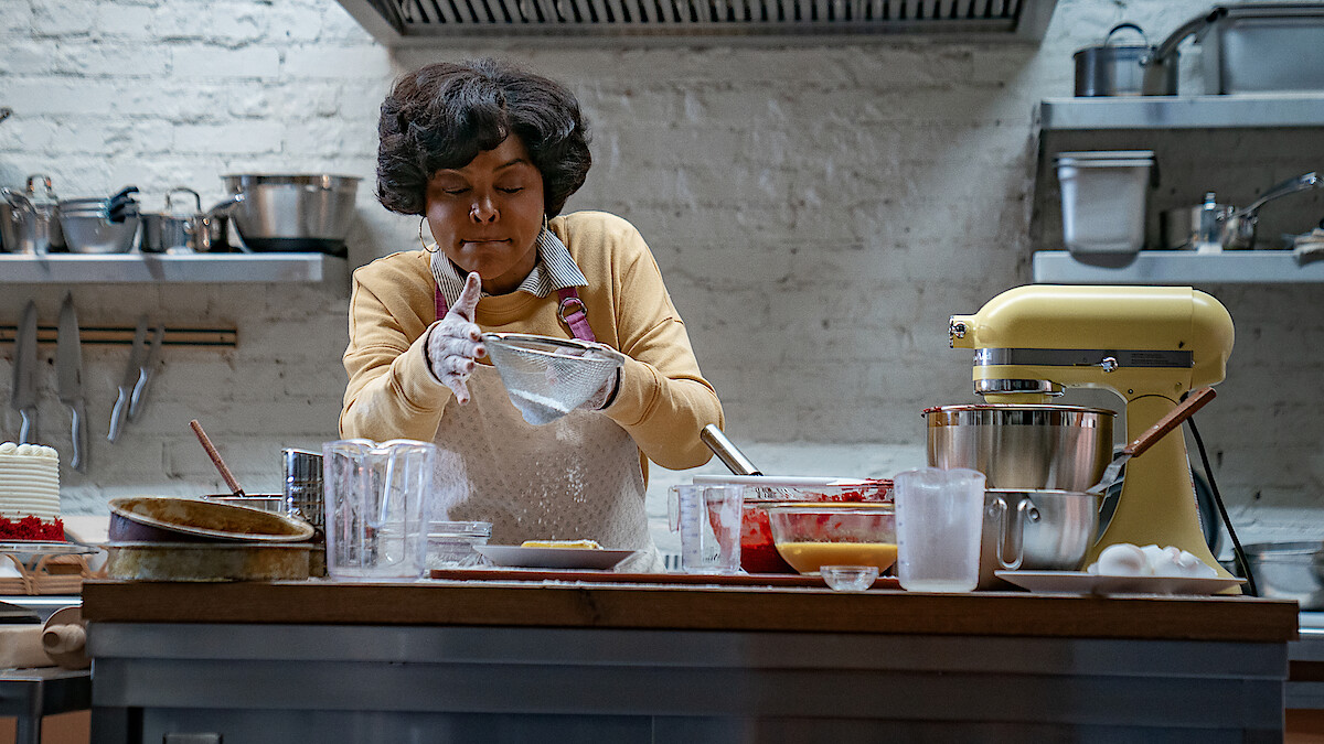 A woman in a kitchen wearing an apron and gloves, concentrating as she prepares food. The kitchen has a stand mixer, various bowls, utensils, and ingredients on a wooden countertop, with shelves and cookware in the background.