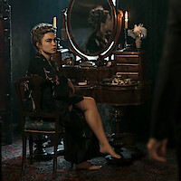 Woman in a dark robe sits at an antique vanity with candles and a mirror, in a dimly lit, elegant room with vintage decor. A blurred figure stands in the foreground, suggesting interaction or tension.