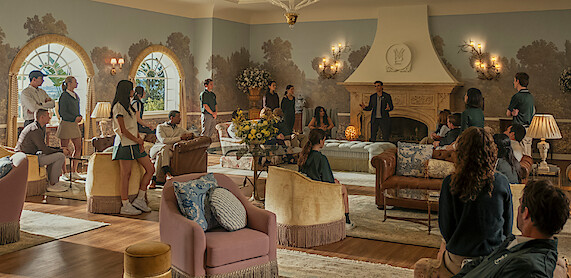 Group of students and adults gathered in a spacious, elegant living room with large windows, ornate fireplace, chandeliers, and stylish furniture, appearing to listen to a speaker in a formal or school setting.