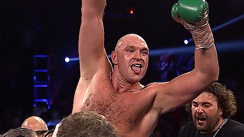 Tyson Fury in a ring raising his arm in victory, wearing green gloves and celebrating with his team under bright arena lights, surrounded by cheering people.