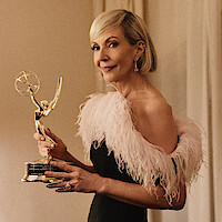 Woman in an elegant dress with feather details stands indoors holding an Emmy award, smiling, with neutral-colored curtains in the background and warm, soft lighting.