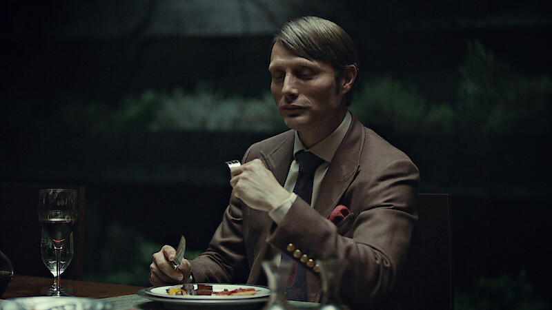 A well-dressed man sits alone at a dimly lit dinner table with a formal place setting, holding a fork and knife, ready to eat. The background is dark and blurred, suggesting an elegant, intimate indoor setting.