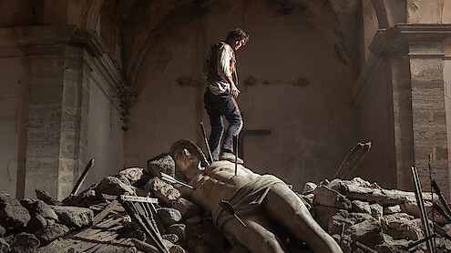 A person stands on rubble inside a damaged, old stone building, with a large broken statue lying among the debris, creating a dramatic and somber atmosphere.