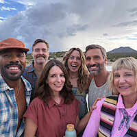 Six smiling adults taking a group selfie outdoors with mountains, desert, and cloudy sky in the background. They appear happy and relaxed, dressed in casual clothing, enjoying a scenic nature outing together.