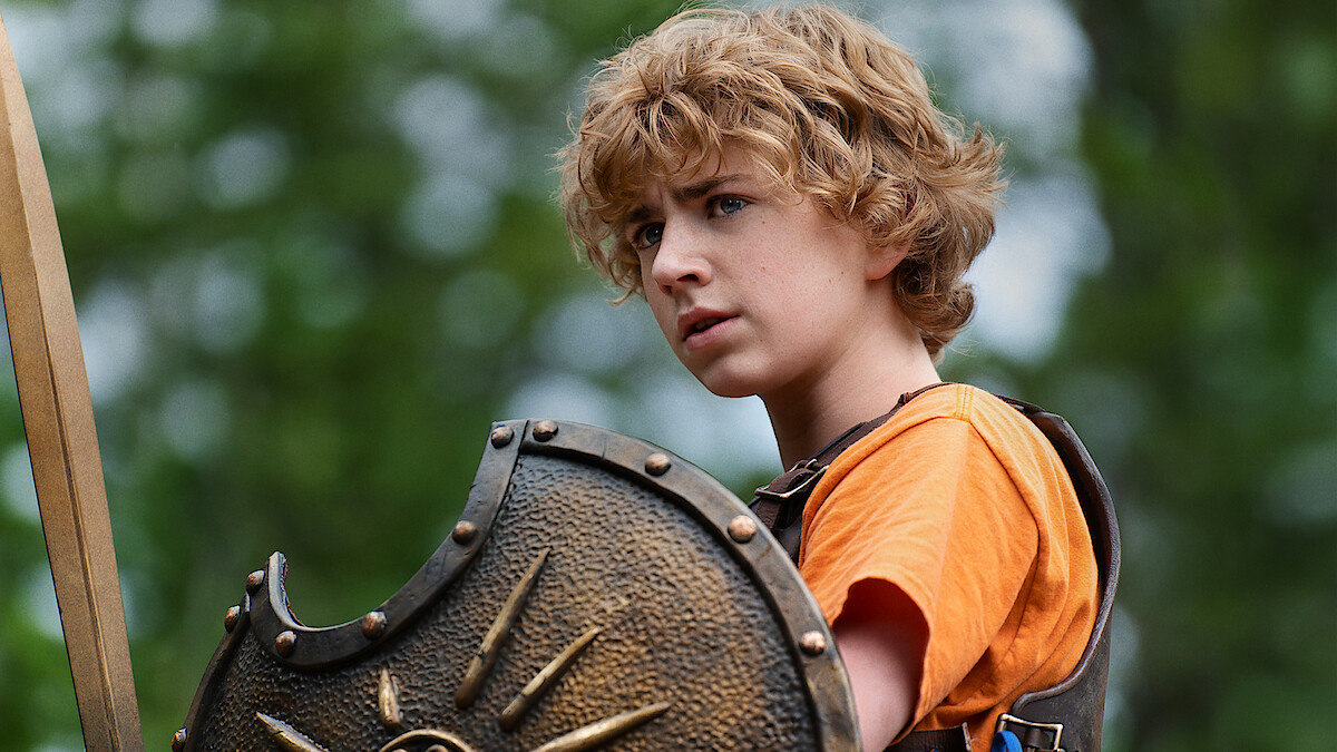 A young person with curly blonde hair in an orange shirt and armor holds a decorated shield outdoors, surrounded by green trees and natural light, appearing alert and determined.