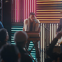 Musician wearing a fur vest plays keyboard and sings on stage, with colorful neon lights in the background and an audience watching the performance in a lively indoor concert setting.
