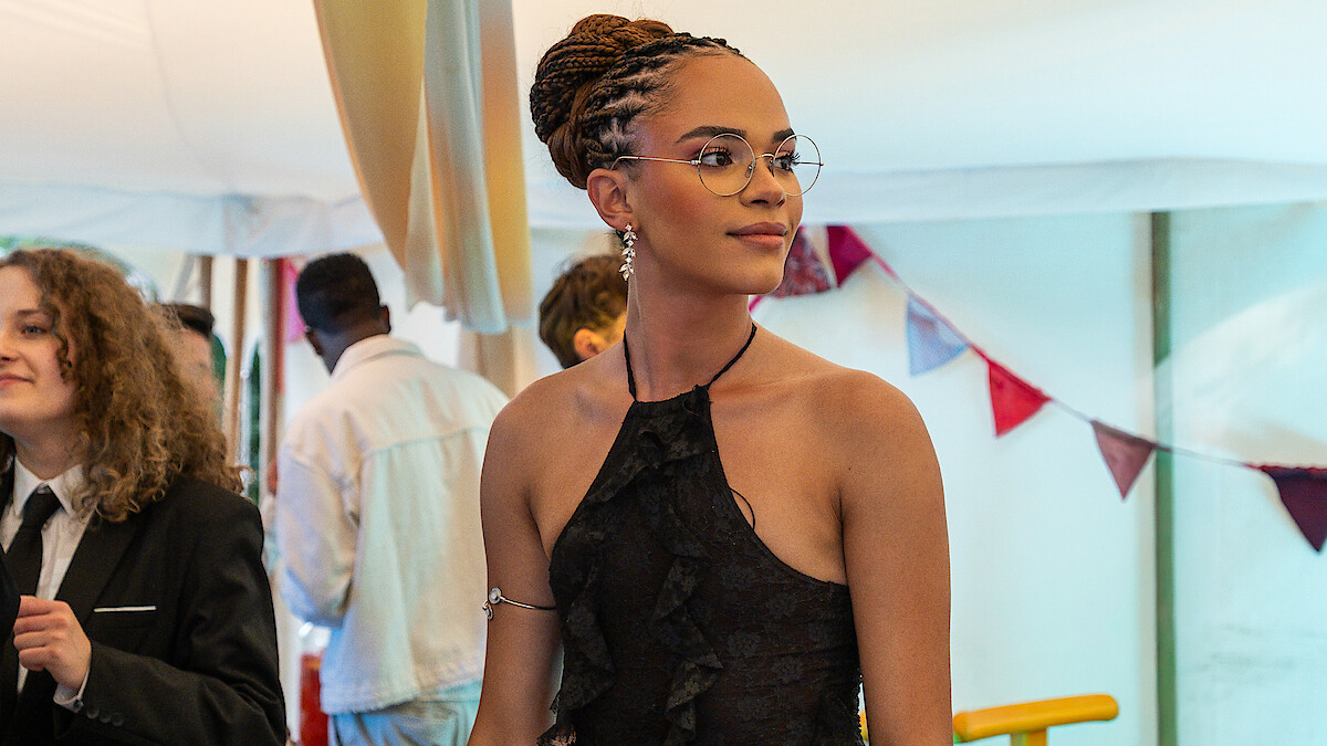 Yasmin Finny in a black dress with braided hair and glasses stands at a festive indoor event with bunting and other guests in formal attire, creating a celebratory atmosphere.