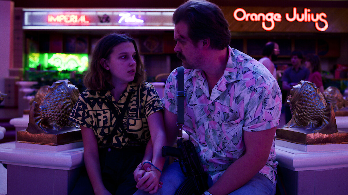 A man and a teenage girl sit closely on a mall bench, surrounded by neon lights and storefronts.