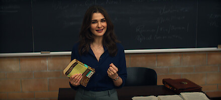 Rachel Weisz as The Protagonist standing in front of classroom chalkboard, holding book and smiling, with desk in foreground holding papers and a briefcase, academic setting.