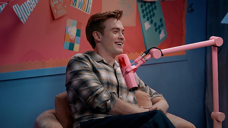 Kit Connor sitting and talking into a pink microphone in a colorful studio with artwork and notes on the wall behind them.