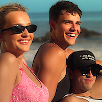 Three young adults relaxing on a sunny beach with rocks in the background, smiling and wearing sunglasses and casual beachwear.