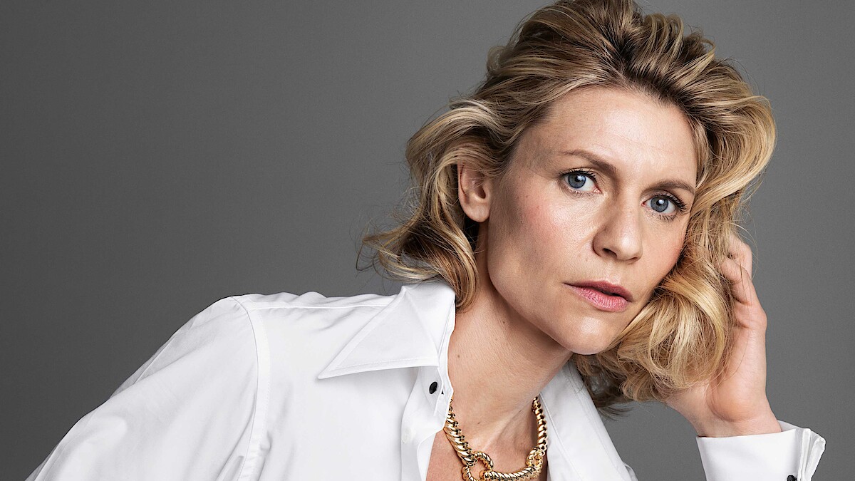 Woman with wavy blonde hair in a white shirt and gold chain necklace, posed against a plain grey background, looking directly at the camera with a thoughtful and serious expression.