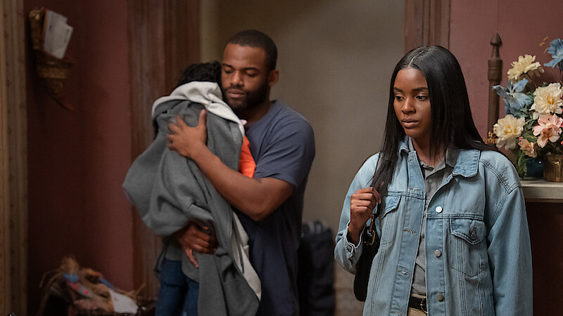 A man holding a child stands beside a woman in a denim jacket. They are indoors in a warmly lit room with flowers on a shelf and decorative objects in the background. The mood appears serious or contemplative.