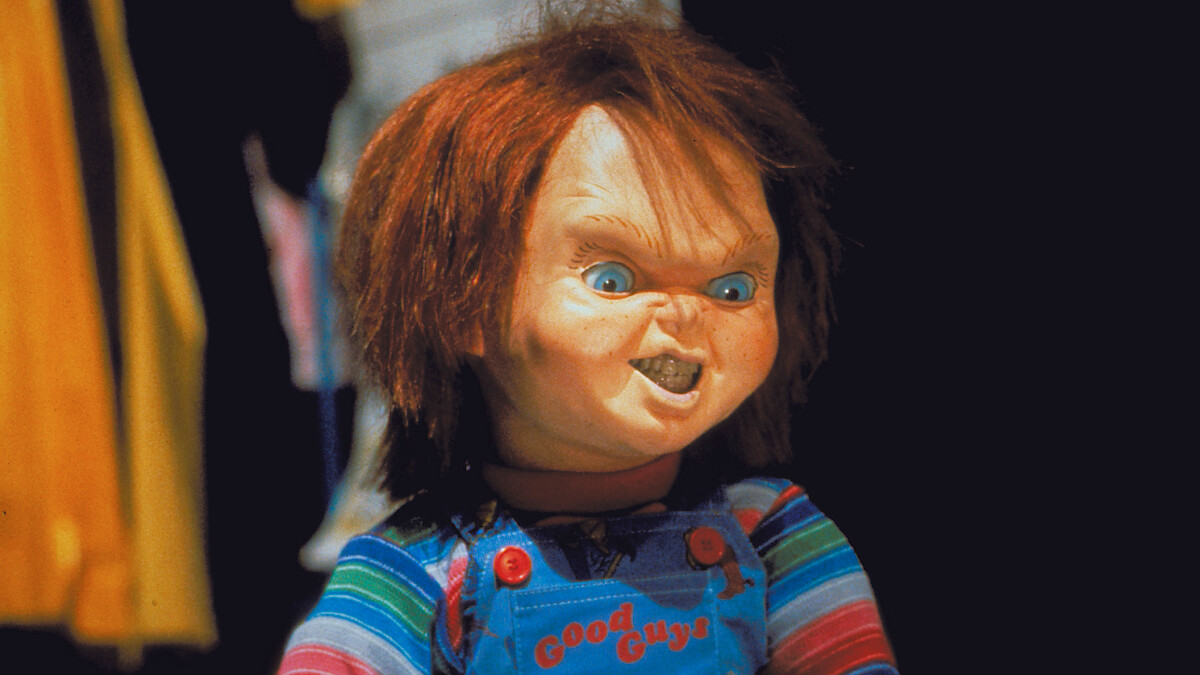 A creepy doll with red hair and overalls stands indoors, wearing a striped shirt. The background is dark with indistinct, colorful objects and clothing visible. The doll has a menacing facial expression.