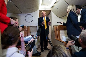 A man in a suit speaks to a group of reporters holding microphones in the aisle of a private jet, with cream-colored seats and overhead lighting.