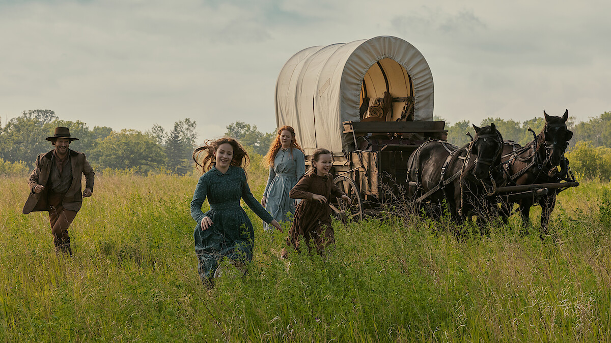Four people in pioneer-era clothing run through a grassy field beside a covered wagon pulled by two horses, with trees and sky in the background, evoking a historical or frontier setting.