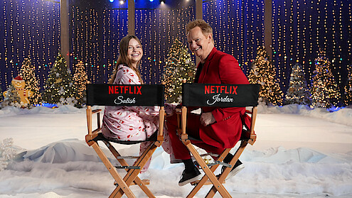Two people smiling while sitting on director’s chairs with Netflix logos in a festive winter set decorated with snow, Christmas trees, twinkling lights, and holiday decorations, creating a cheerful holiday atmosphere.