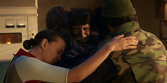 Eleven, Jonathan Byers, Will Byers, and Mike Wheeler sharing an emotional group hug indoors, with eyes closed, in a warmly lit room.