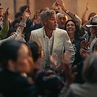 A man in a white suit stands in the center of a lively crowd, surrounded by people celebrating with raised hands and joyful expressions in a festive atmosphere.