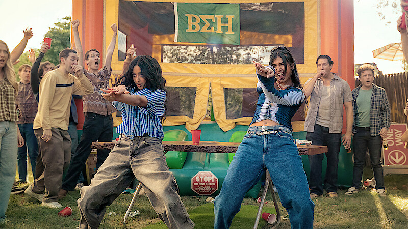 Two women dance energetically at an outdoor party with a crowd of cheering people, in front of a colorful bouncy house decorated with Greek letters, on a sunny day.